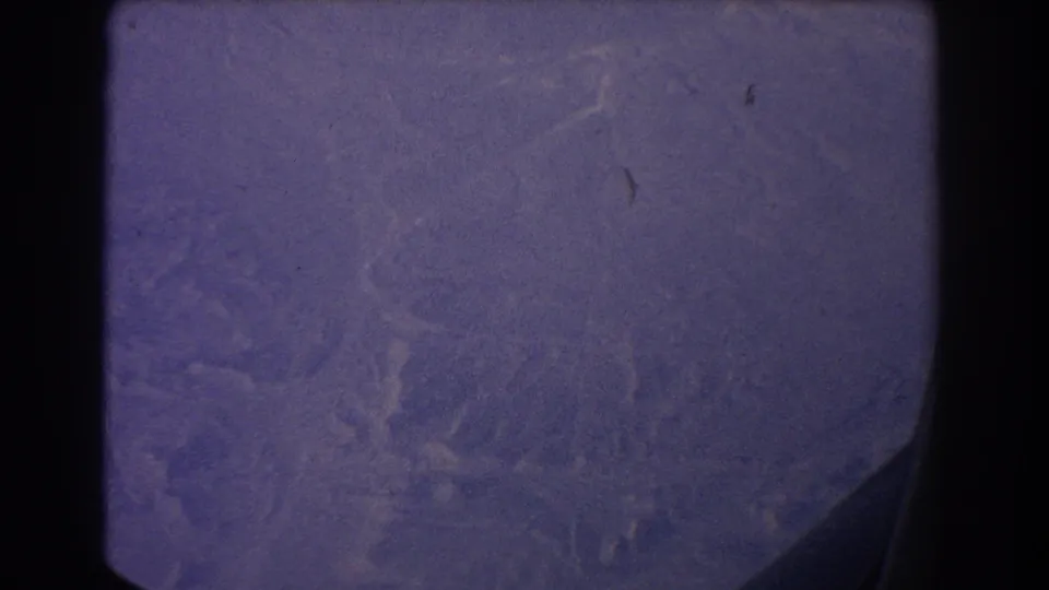 1967 Sydney-area 8mm frame with a hazy aerial view of rugged coastal terrain, rounded gate edges, visible scratches, and a dark lower-right shape.