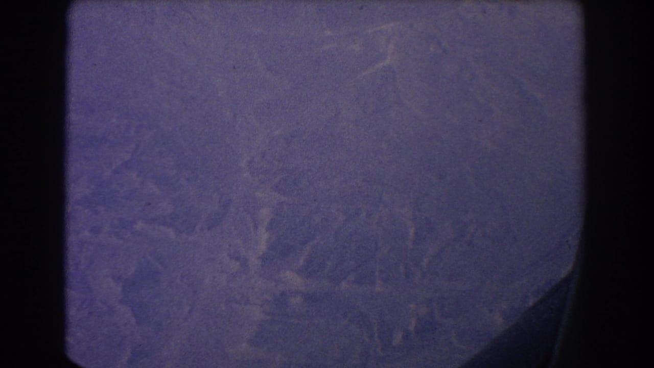 Purple-tinted aerial terrain seen through an airplane window with dark film borders.