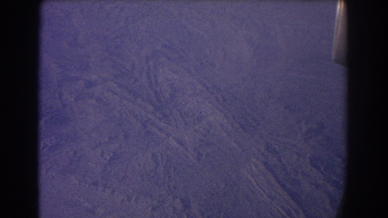 Purple-tinged arid terrain seen through an airplane window with dark film borders.