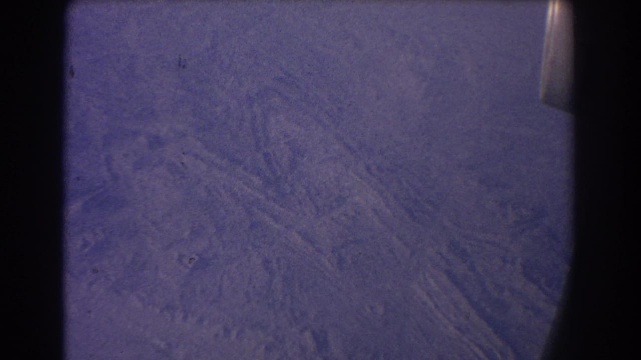 Arid landscape with faint linear patterns and aircraft-window edge at the right.