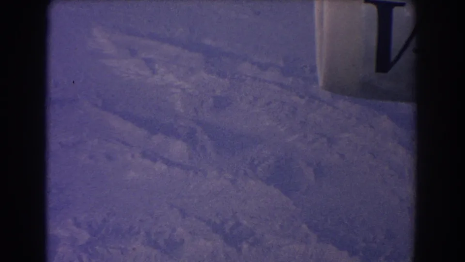 Snowy terrain remains visible below, with an aircraft window or frame element on the right and 8mm grain across the image.