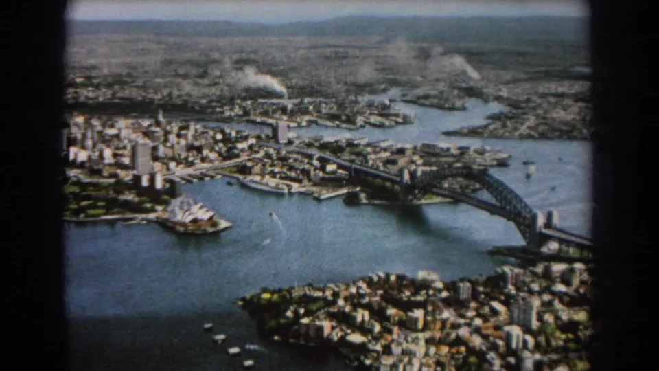 1967 8mm aerial frame of Sydney Harbour, showing the Harbour Bridge, Opera House, waterfront development, blue water, and boats below.