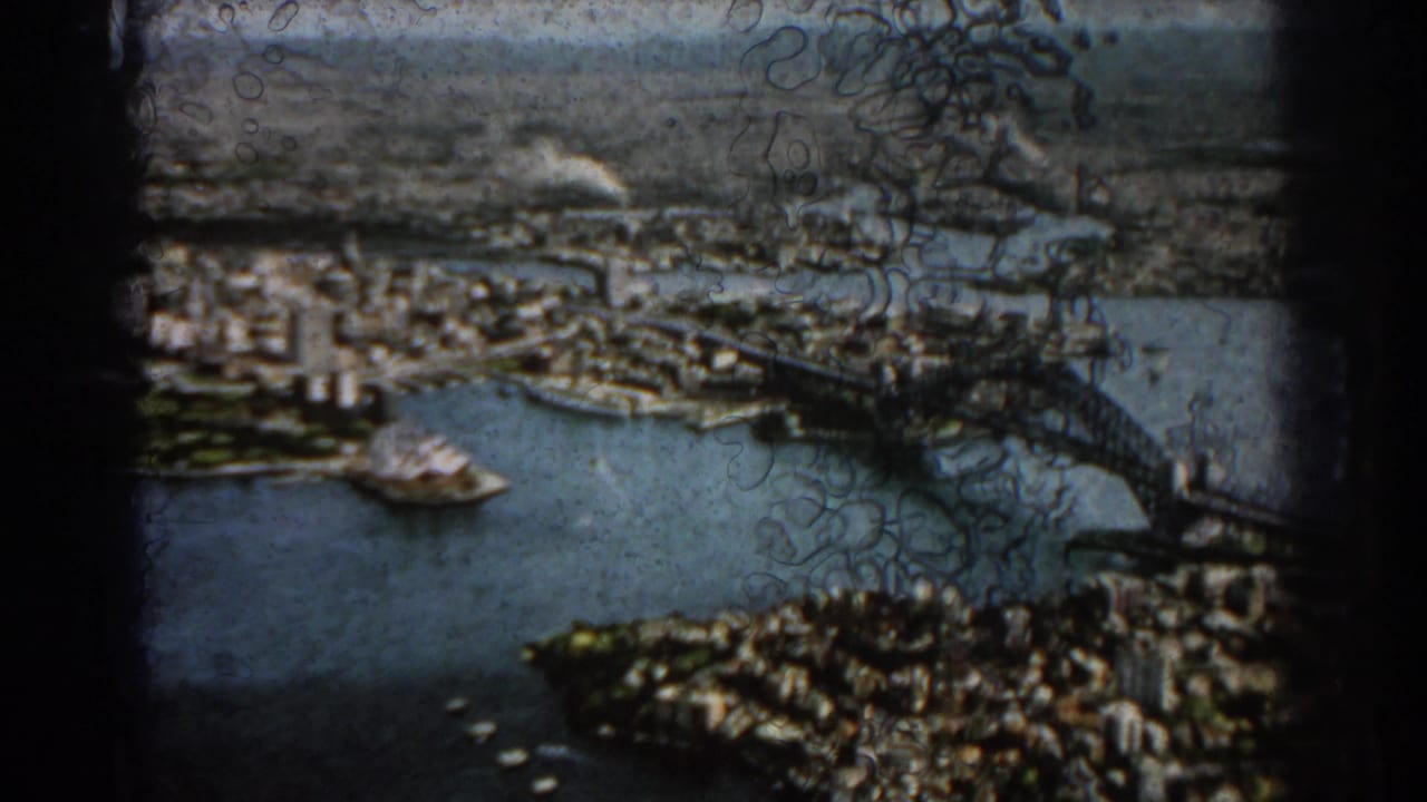 1967 8mm aerial view over Sydney Harbour, showing the Harbour Bridge, waterfront, and dense city blocks.