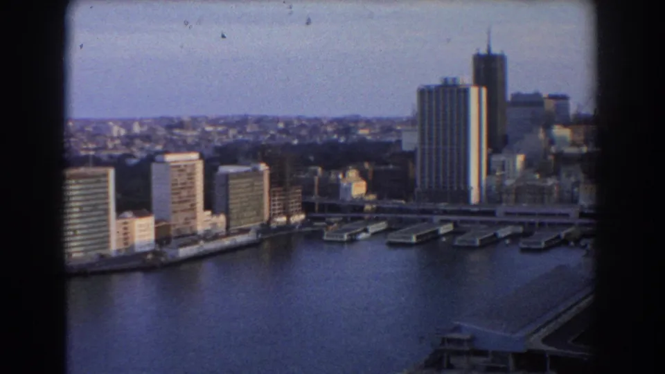 Sydney harbor in 1967 from a high vantage point, with mid-century towers, wharf structures, calm water, and softened home-movie detail.