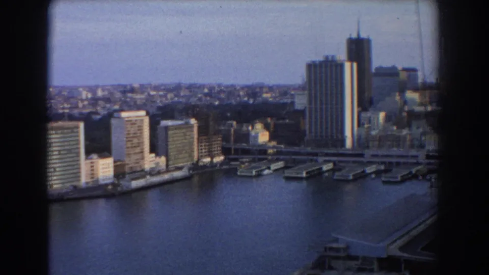 Late-1960s Sydney harbor skyline view with a prominent tower, waterfront buildings, piers, calm blue-gray water, and 8mm scan texture.