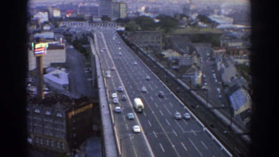 Soft 8mm frame of 1967 Sydney highway traffic, with a white bus, several cars, nearby buildings, and a visible commercial sign.
