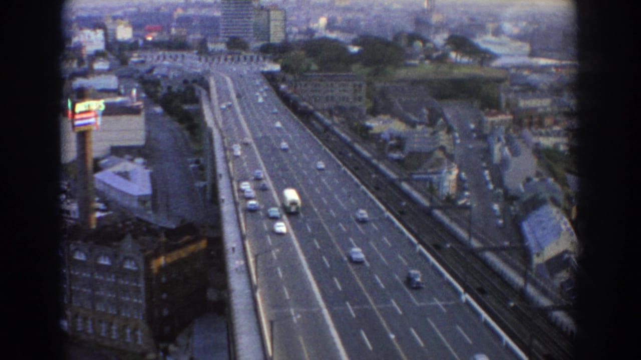 1967 Sydney home-movie frame showing traffic on a broad urban road from a high vantage, with buildings and skyline in soft focus.