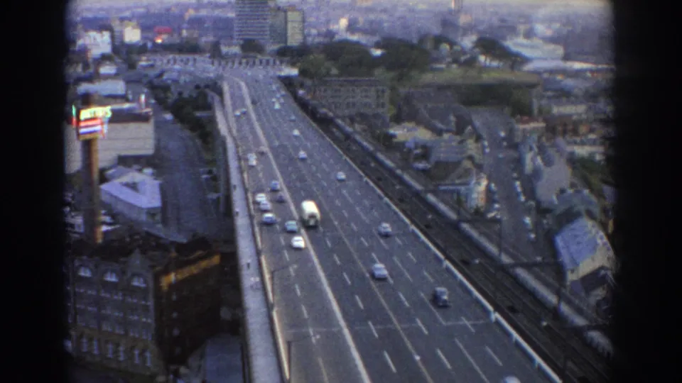 1967 Sydney home-movie frame showing traffic on a broad urban road from a high vantage, with buildings and skyline in soft focus.