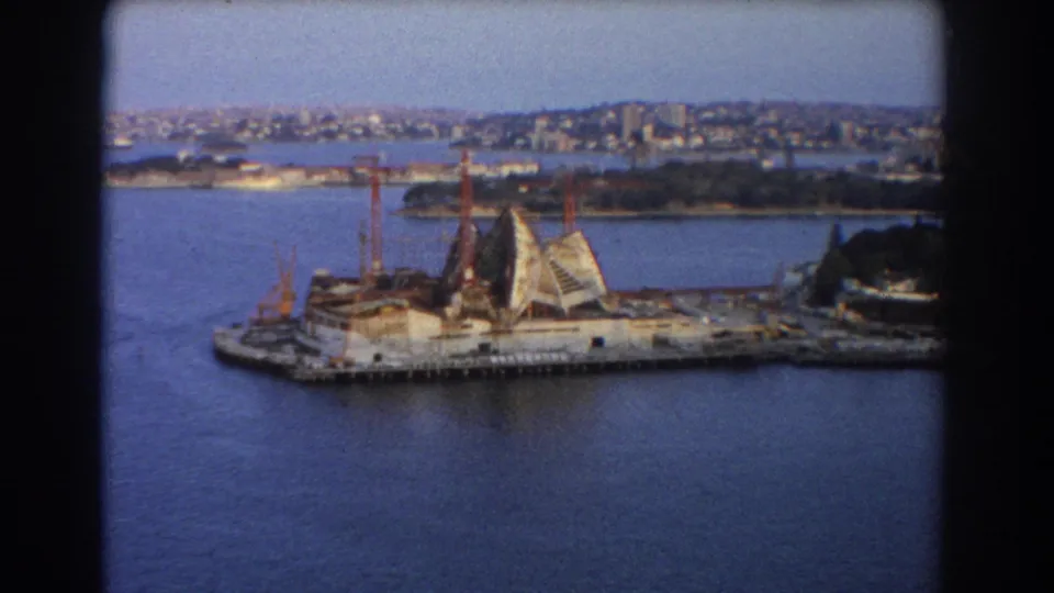 1967 Sydney harbor frame showing the Opera House under construction, with cranes, shell forms, water, and distant shoreline.