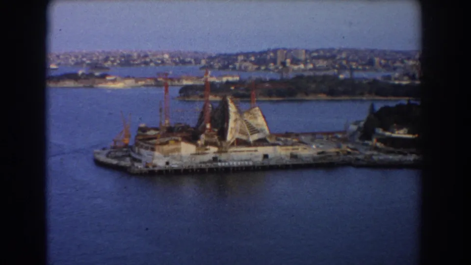 Sydney Opera House construction seen from above in 1967, with cranes over the harbor site and buildings across the water.