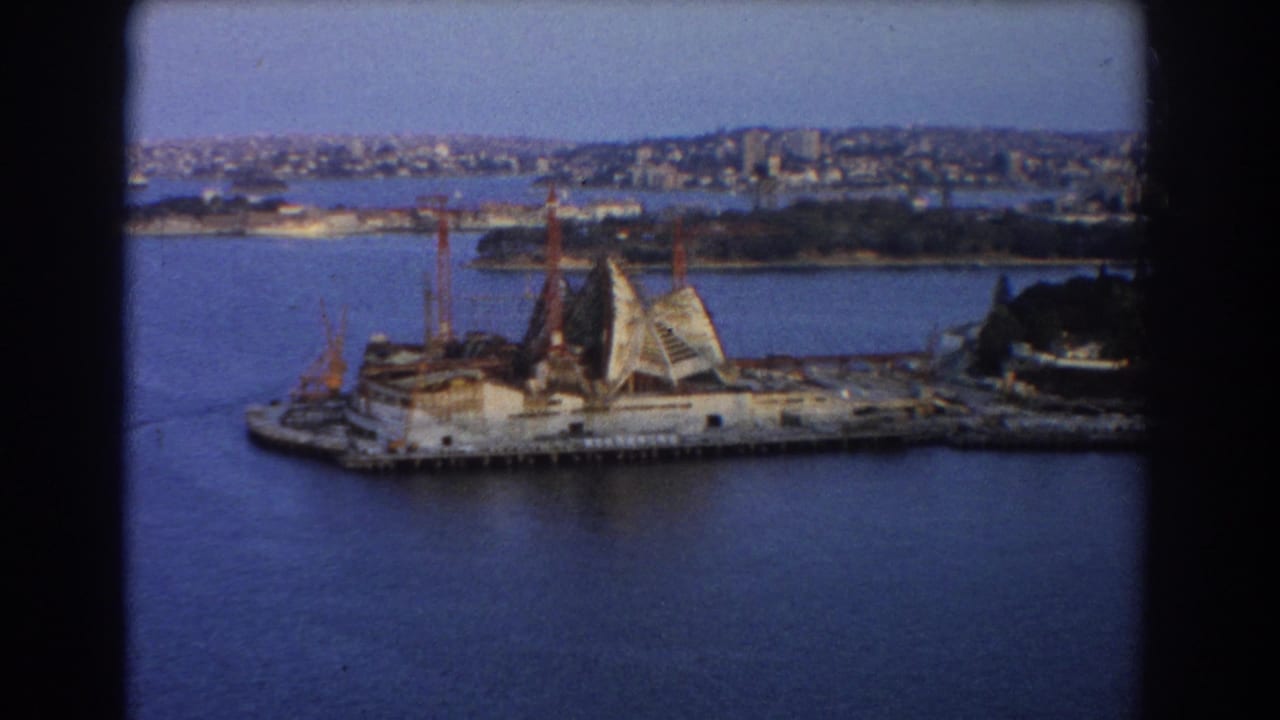 Harbour view of the Opera House construction site with cranes and city skyline.