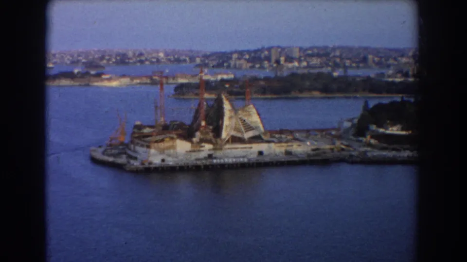 1967 Sydney view across blue harbour water toward the unfinished Opera House, surrounding cranes, waterfront, and distant city buildings.