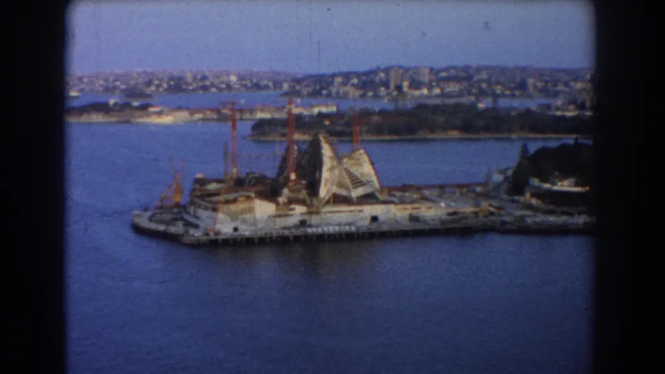 Home-movie frame from Sydney in 1967 showing the Opera House construction area, cranes, harbour water, and city skyline beyond.