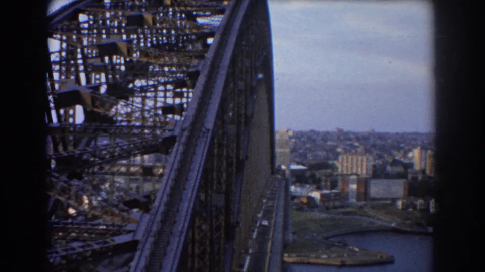 1967 view from Sydney Harbour Bridge, looking along steel bridge structure toward roadway, harbour water, and city buildings below.