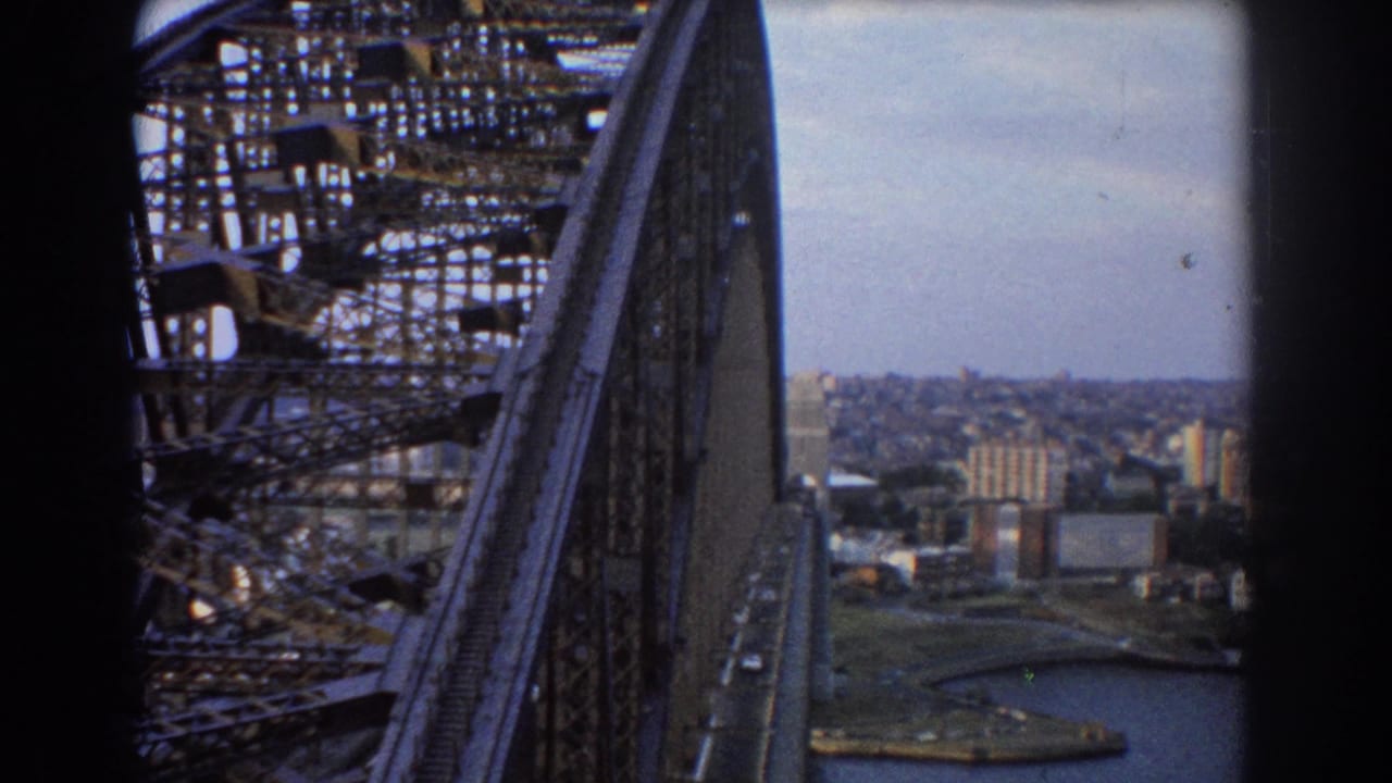 High-angle view along Sydney Harbour Bridge with roadway, truss steel, harbor water, and distant city buildings.