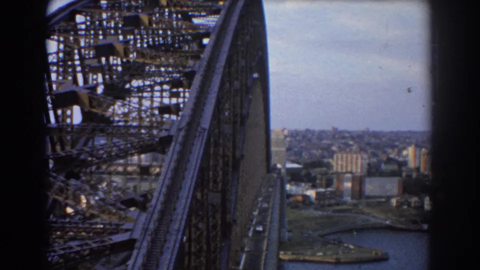 1967 8mm view from Sydney Harbour Bridge showing steel truss work, a road lane, harbor water, and the city beyond.