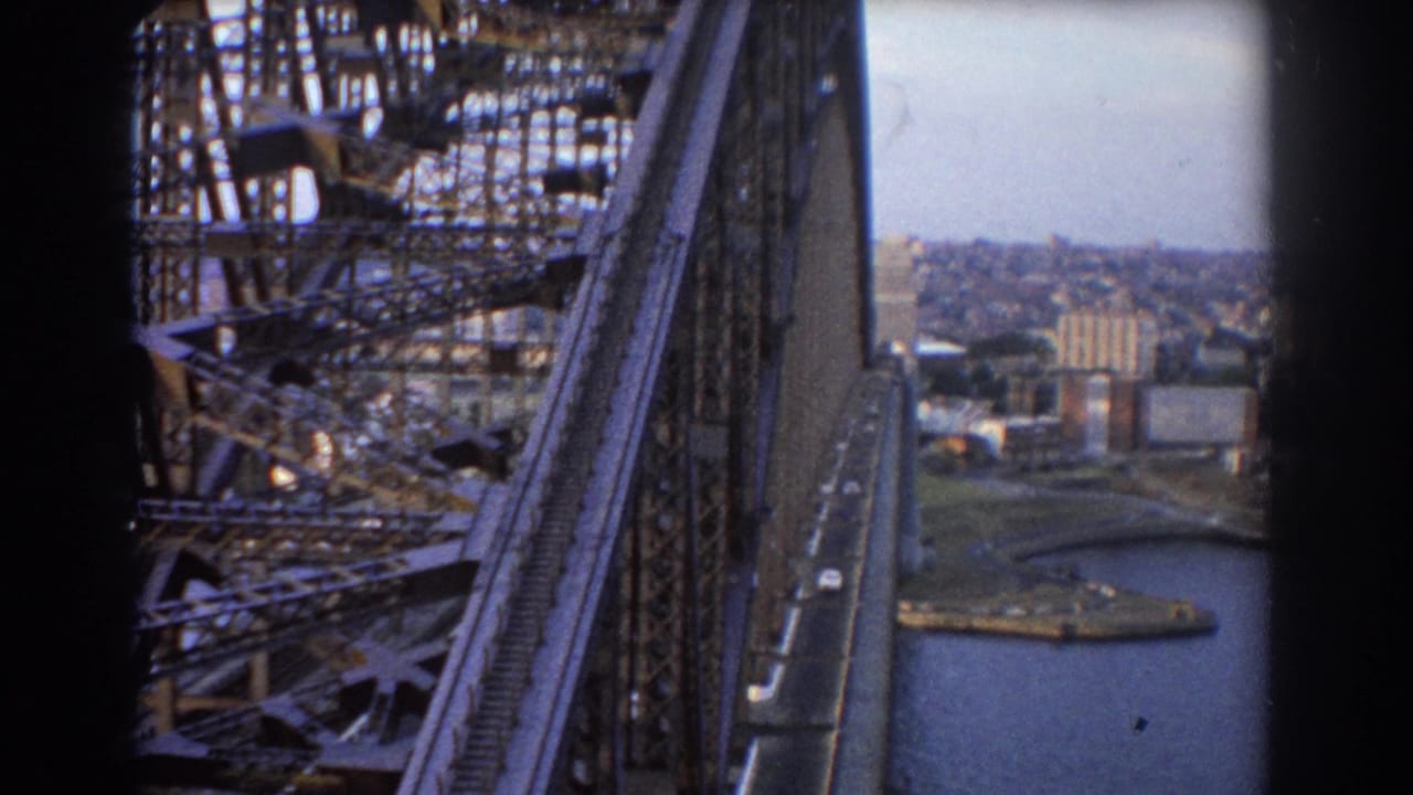 Bridge steelwork and roadway seen from above, with harbor water and city buildings in the background.