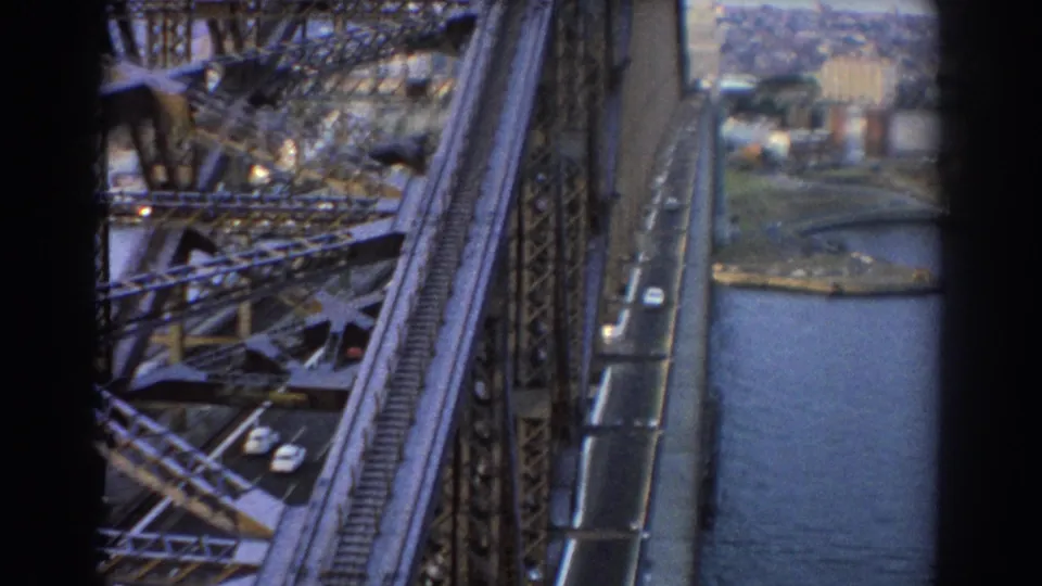 Soft 8mm frame of Sydney Harbour Bridge showing the steel span, road below, harbor shoreline, and city buildings in the distance.