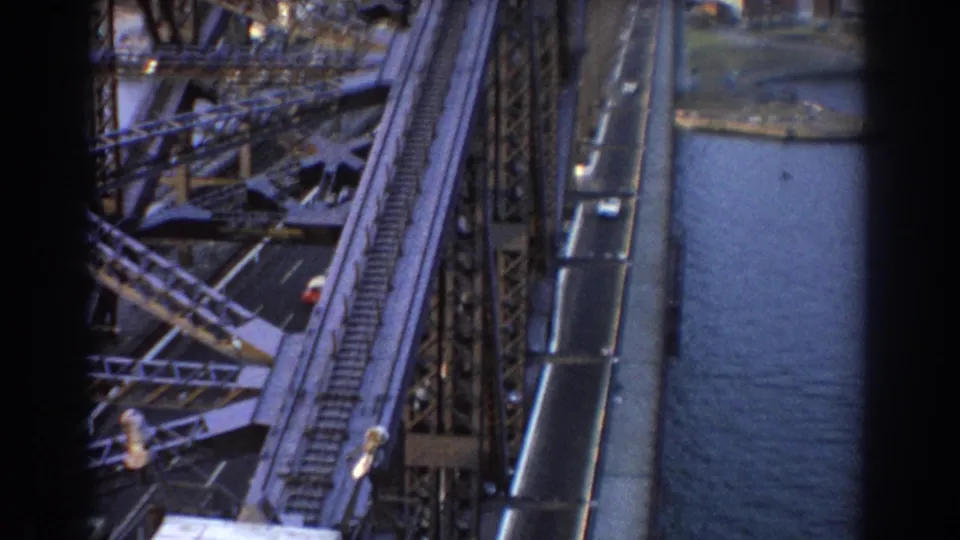 1967 Sydney Harbour Bridge view with steel lattice, roadway traffic, rail corridor area, blue harbor water, and waterfront land below.