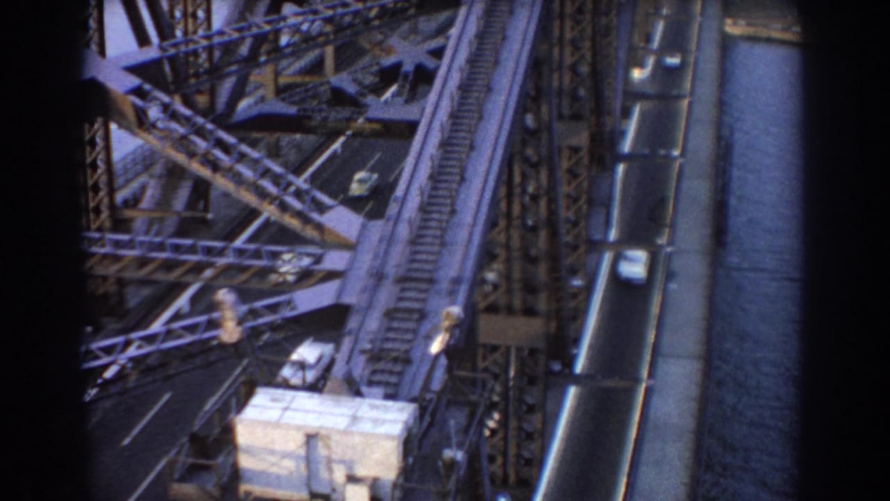 Bridge roadway with cars, steel supports, rail tracks, and water at the side.