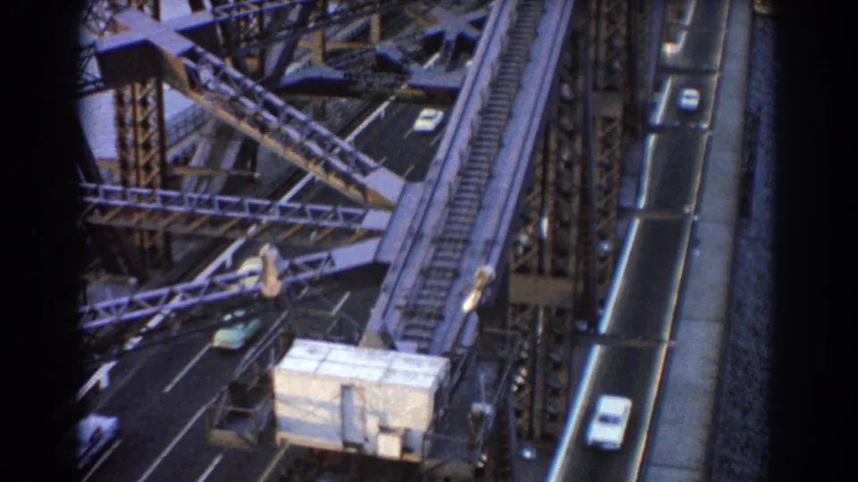 1967 Sydney home-movie frame showing cars on a steel bridge with rail tracks and harbor water nearby.