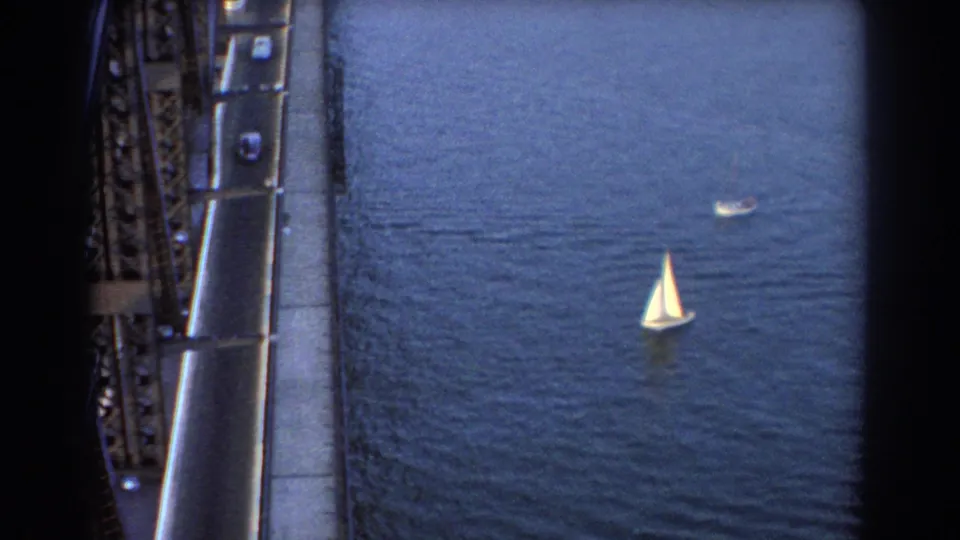 Sydney 1967 bridge traffic frame with open water and a small white sailboat beyond the roadway.