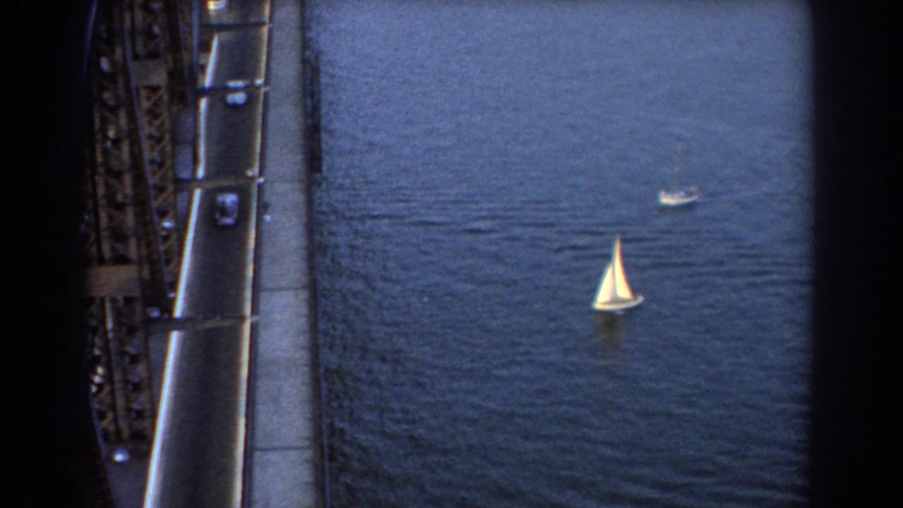 Two sailboats on blue water beside a steel bridge with cars on the roadway.