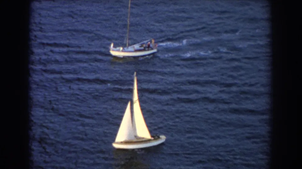 1967 Sydney 8mm frame showing a white-sailed boat in the foreground and a small boat farther back on rippled water.