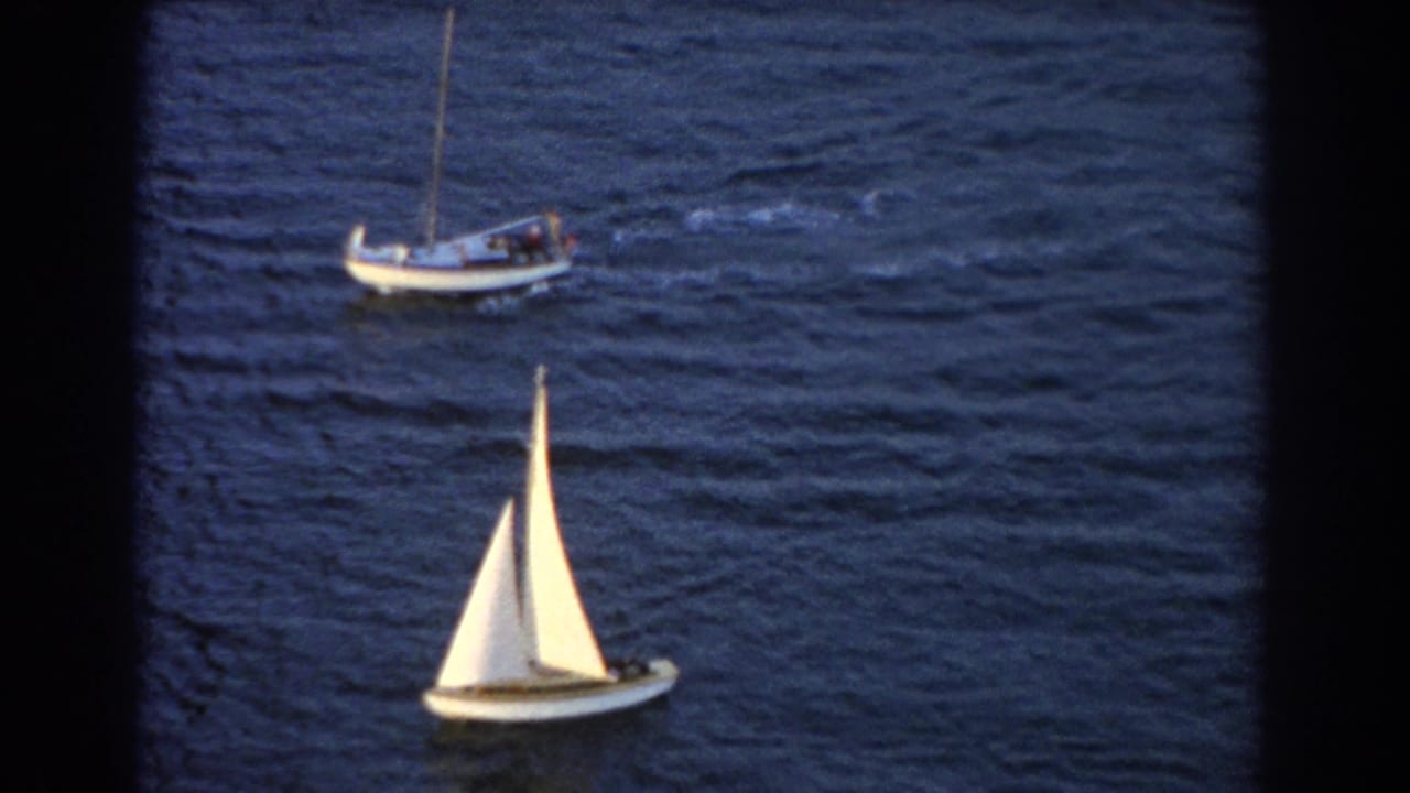 Two small boats on dark blue water, one with white sails in the foreground.