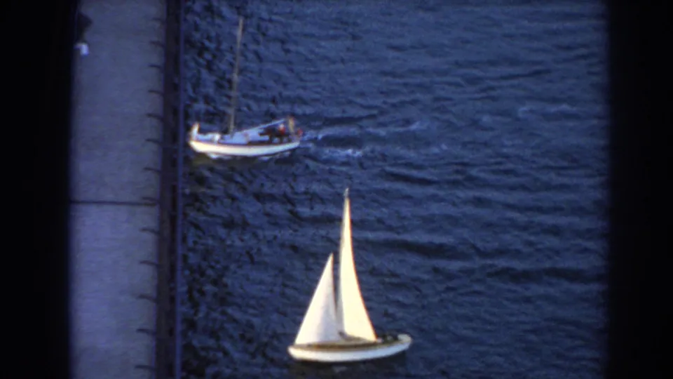 A small white-sailed boat moves through dark blue water below a larger craft in 1967 Sydney-area home-movie footage.