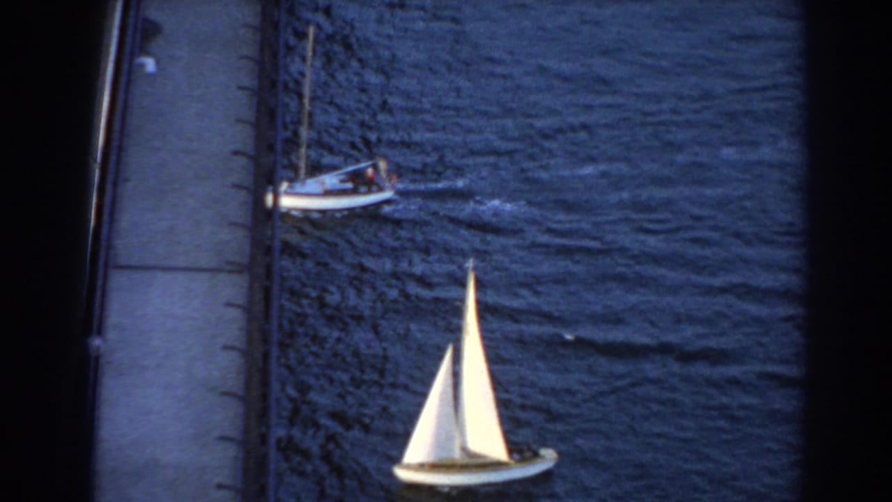 White sailboat below a bridge edge with a small motorboat crossing nearby on dark water.