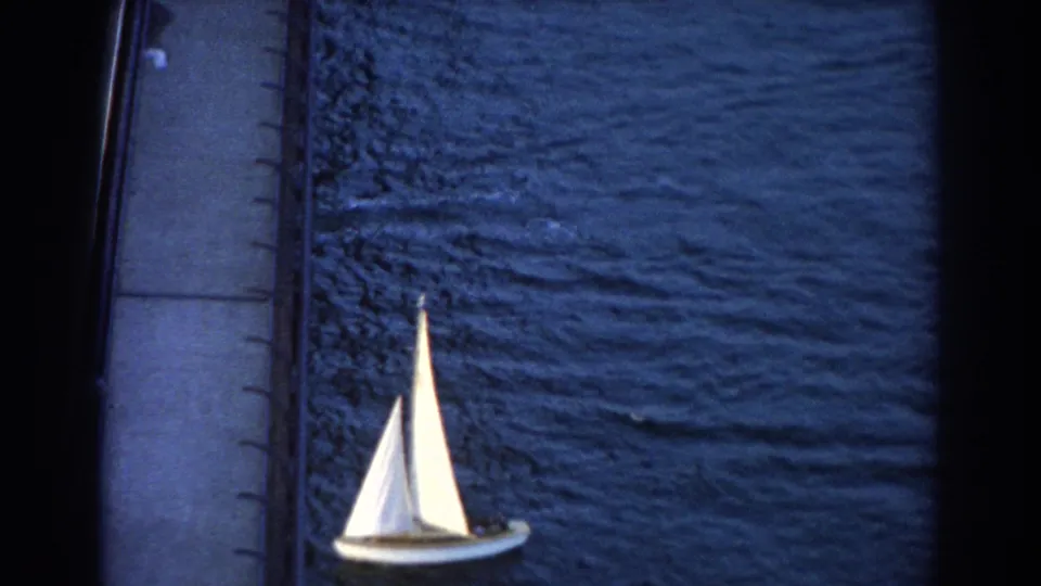 High-angle 1967 Sydney 8mm frame with a white sailboat passing beside a bridge edge over dark rippled harbor water.