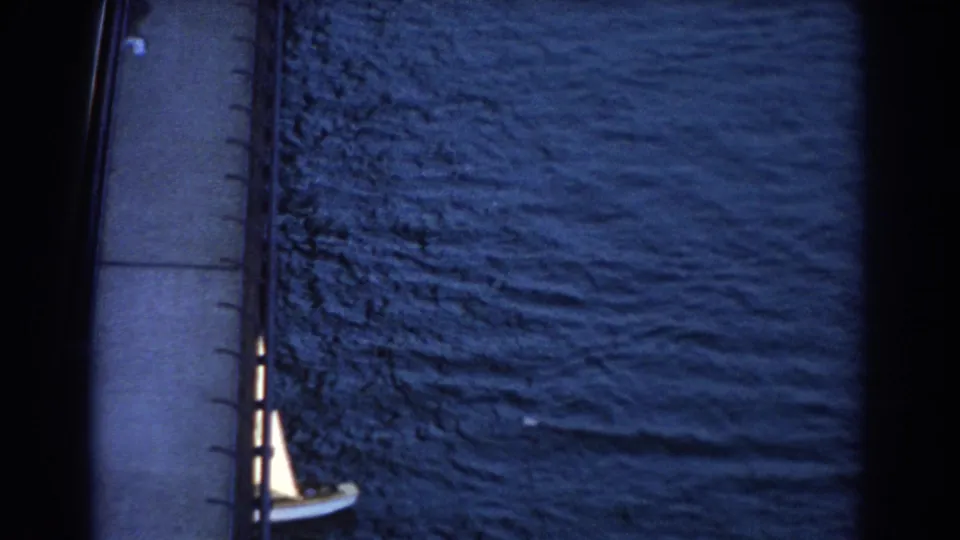 Small sailboat continues under or beside the bridge in 1967 Sydney footage, seen from above against dark blue water.