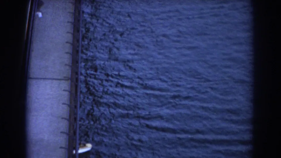High-angle 8mm view in Sydney, 1967, with the sailboat mostly leaving frame beside a concrete bridge and rippled water.