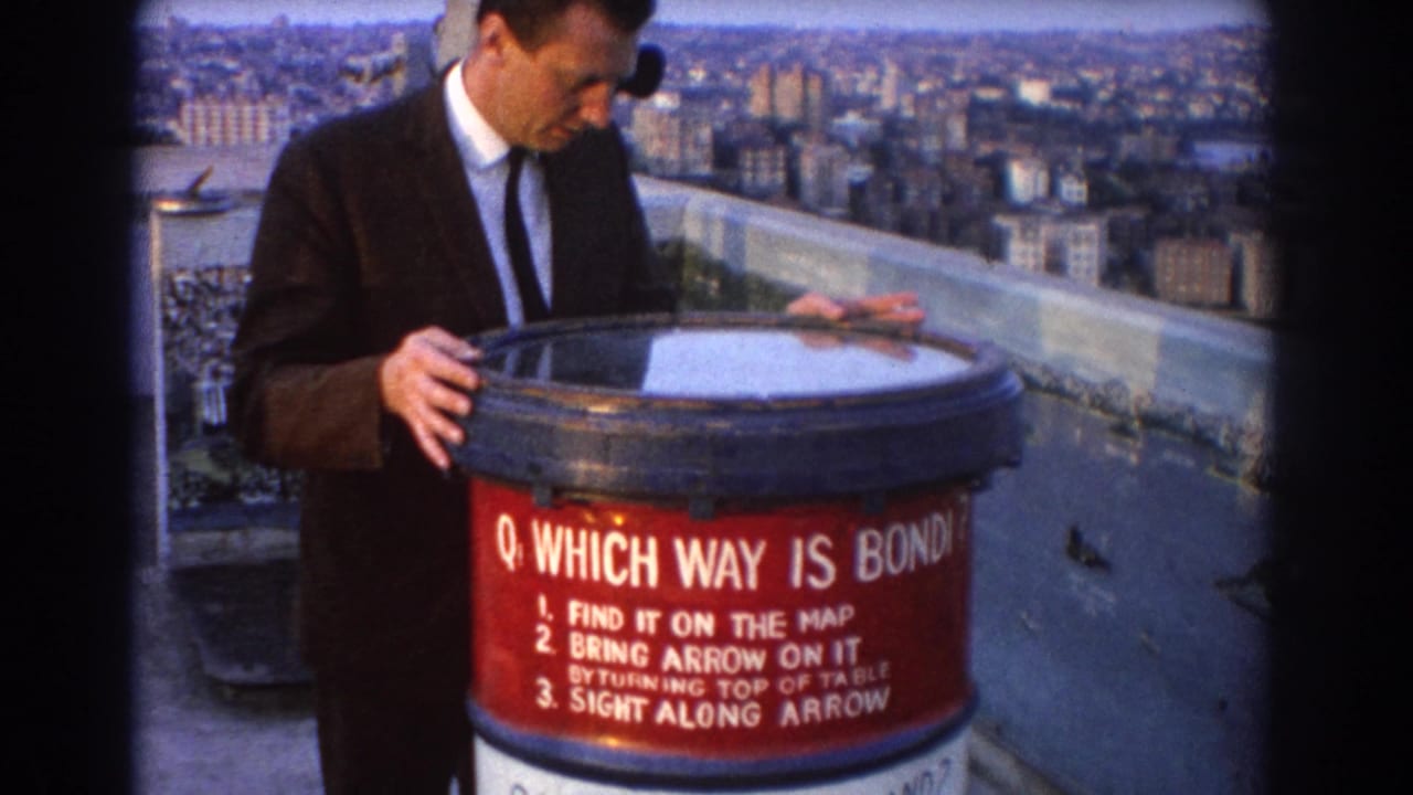 In 1967 Sydney, a suited man uses a red public orientation table at a scenic lookout above the harbor and city skyline.