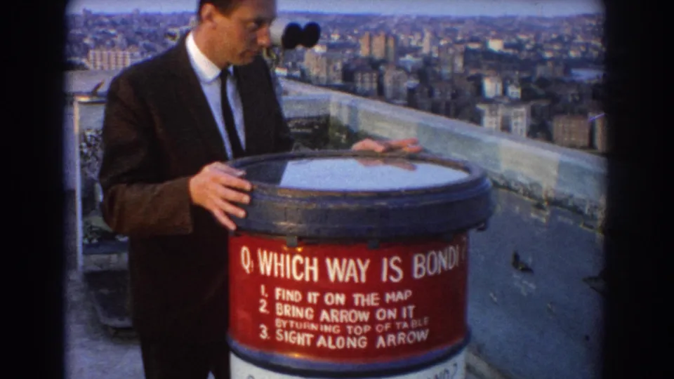 A suited man studies the red orientation table at a Sydney scenic lookout, with harbor water and urban buildings visible beyond.