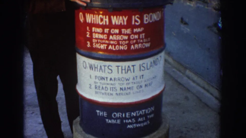 1967 Sydney home-movie frame showing a waterfront orientation sign on a barrel, with a partial adult figure at the left edge.