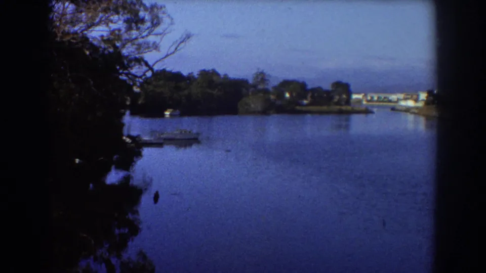 1967 Sydney 8mm view across calm water with a small boat near the shaded shore and low buildings in the distance.