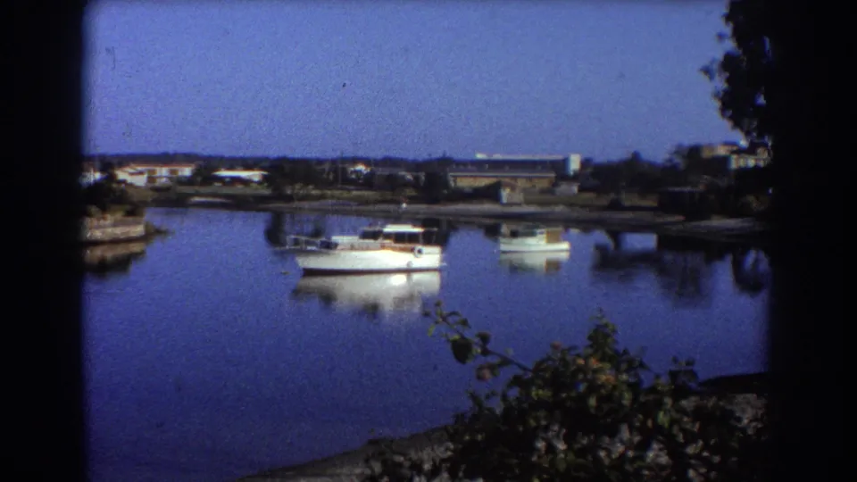 1967 Sydney home-movie frame showing white boats moored on still blue water beneath a low shoreline skyline.