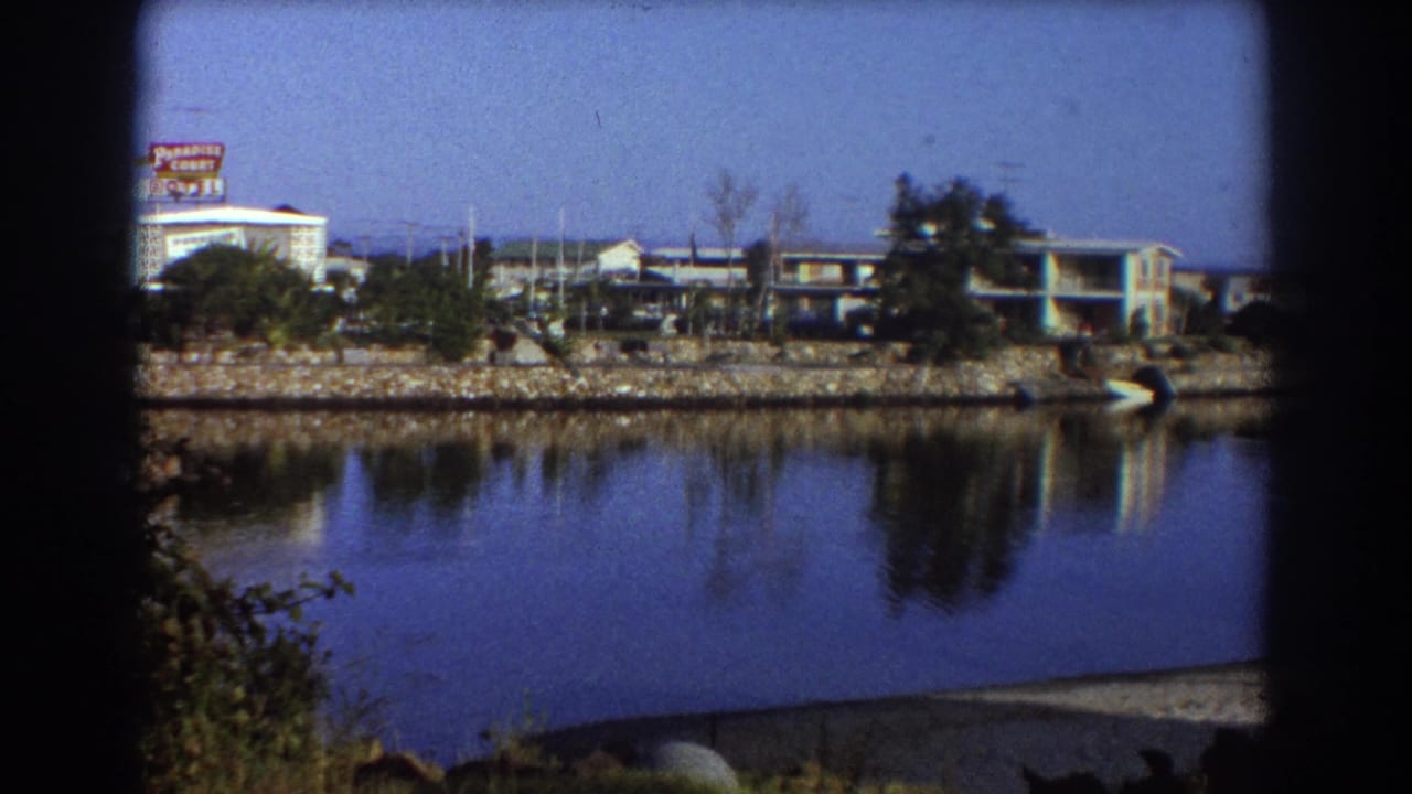 Calm lake with low buildings, a roadside sign, trees, and reflections across the water.