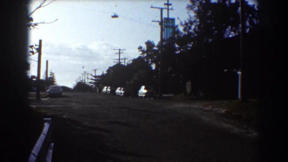 1967 Sydney street view with parked cars, utility poles, dark roadside trees, and a distant industrial structure near the horizon.