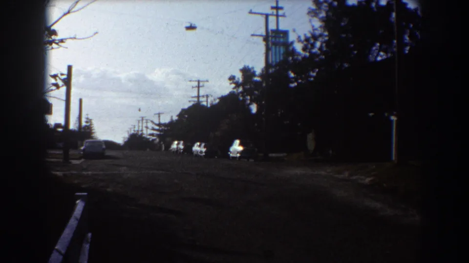 1967 Sydney home-movie street frame showing a parked car, utility poles, roadside trees, and deep shadows along the curb.