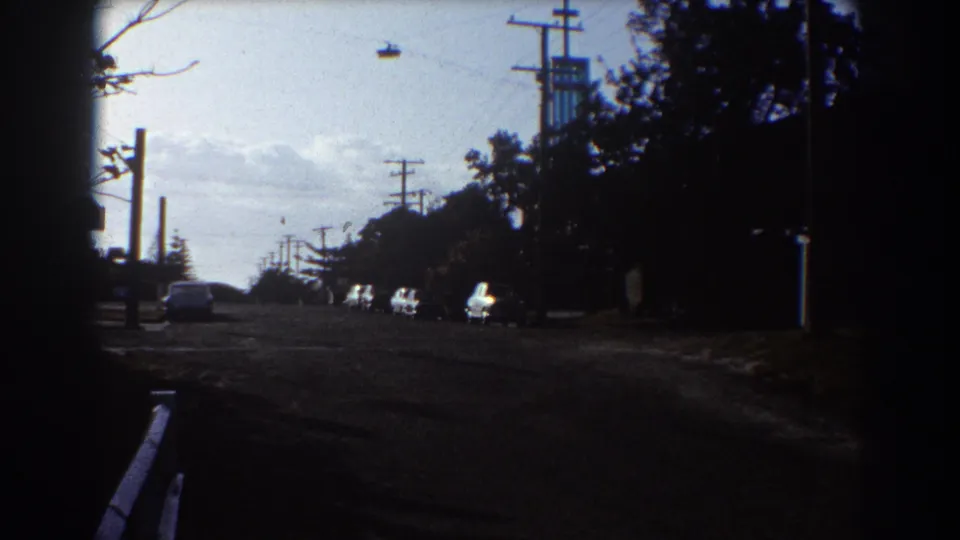1967 Sydney street view with parked cars, utility poles, silhouetted trees, and a small overhead cable or gondola-like object in the sky.