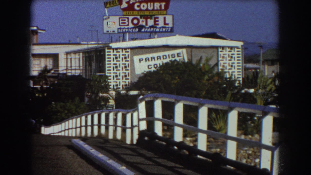 1967 Sydney home-movie frame showing the Paradise Court Motel sign, serviced-apartments lettering, shrubs, and a curved white roadside railing.