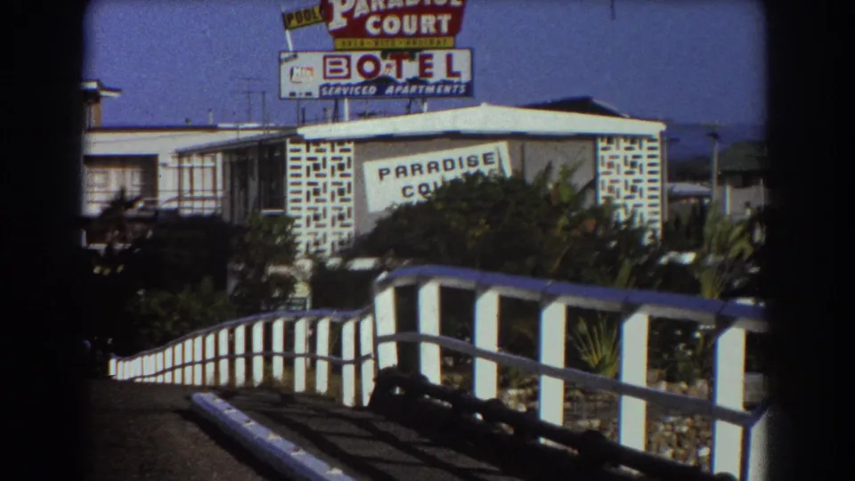 Sydney motel exterior in 1967 with Paradise Court signage, white bridge-like railing, planted shrubs, and low roadside buildings.