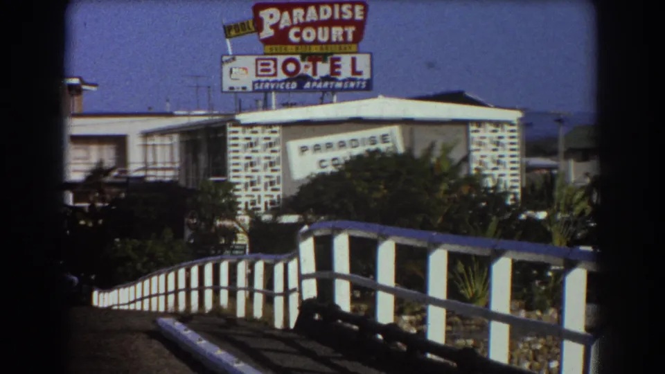 1967 Sydney frame with the Paradise Court Motel sign partly framed by shrubs, a low building, and a curved white railing in the foreground.