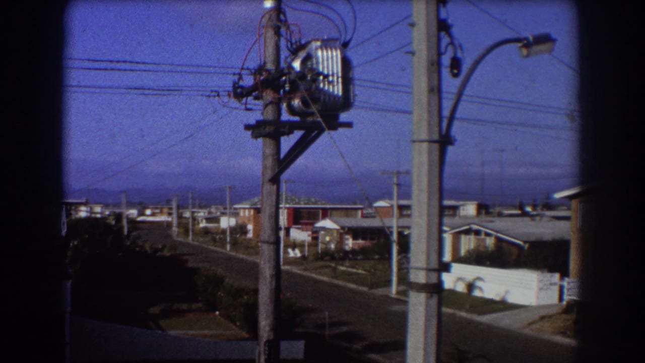 Suburban street seen past utility poles and overhead wires under a blue sky.