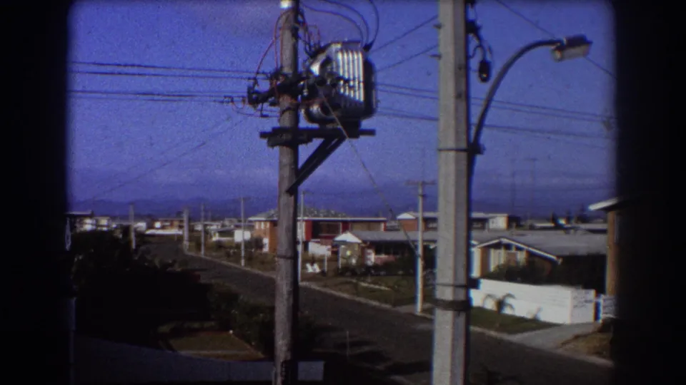 1967 8mm view of an Australian suburban street with wooden utility poles, transformer equipment, wires, and low houses.