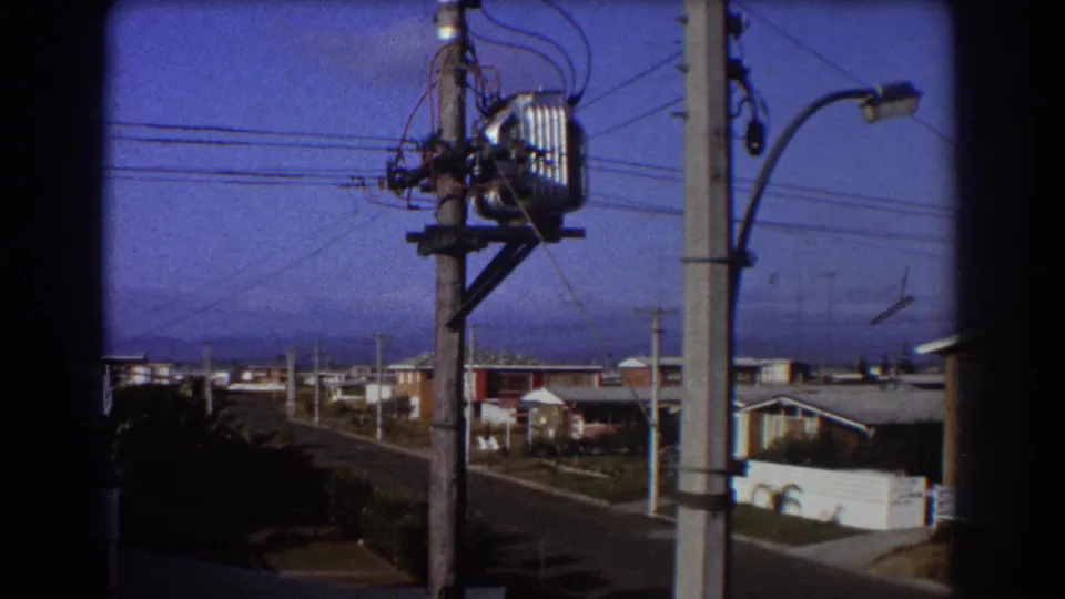 1967 home-movie frame showing suburban Australian houses beyond utility poles, transformer hardware, wires, and a curved streetlight.