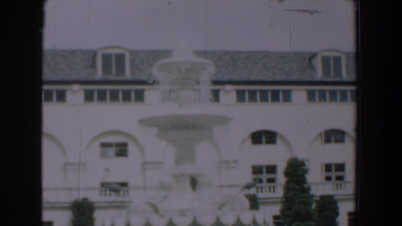 1957 Florida home-movie frame showing a large white building with arched windows, roof dormers, a fountain, and shrubs.
